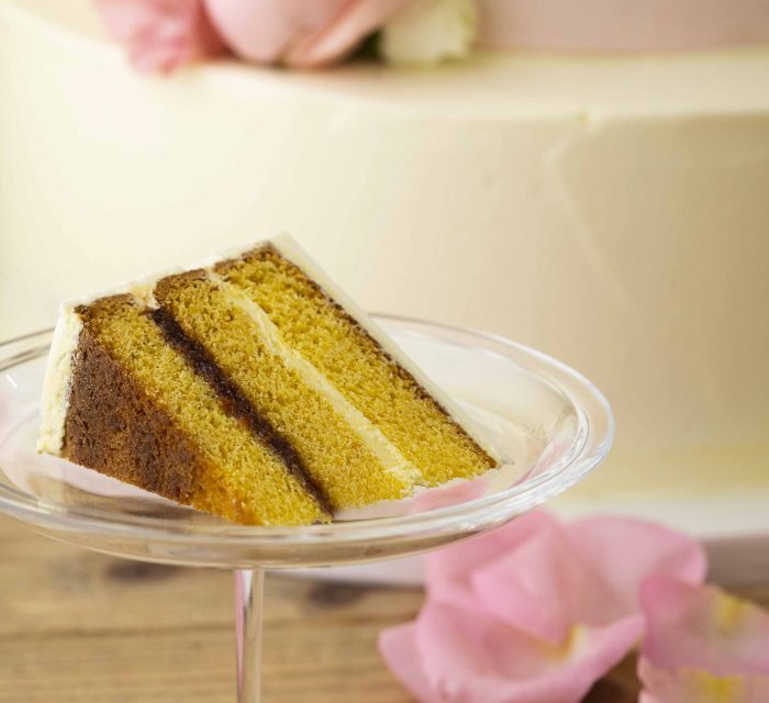 pezzo di torta nuziale servito su un alzatina di vetro. Sullo sfondo, su un tavolo, dei petali di rosa e una torta nuziale con una rosa rosa su